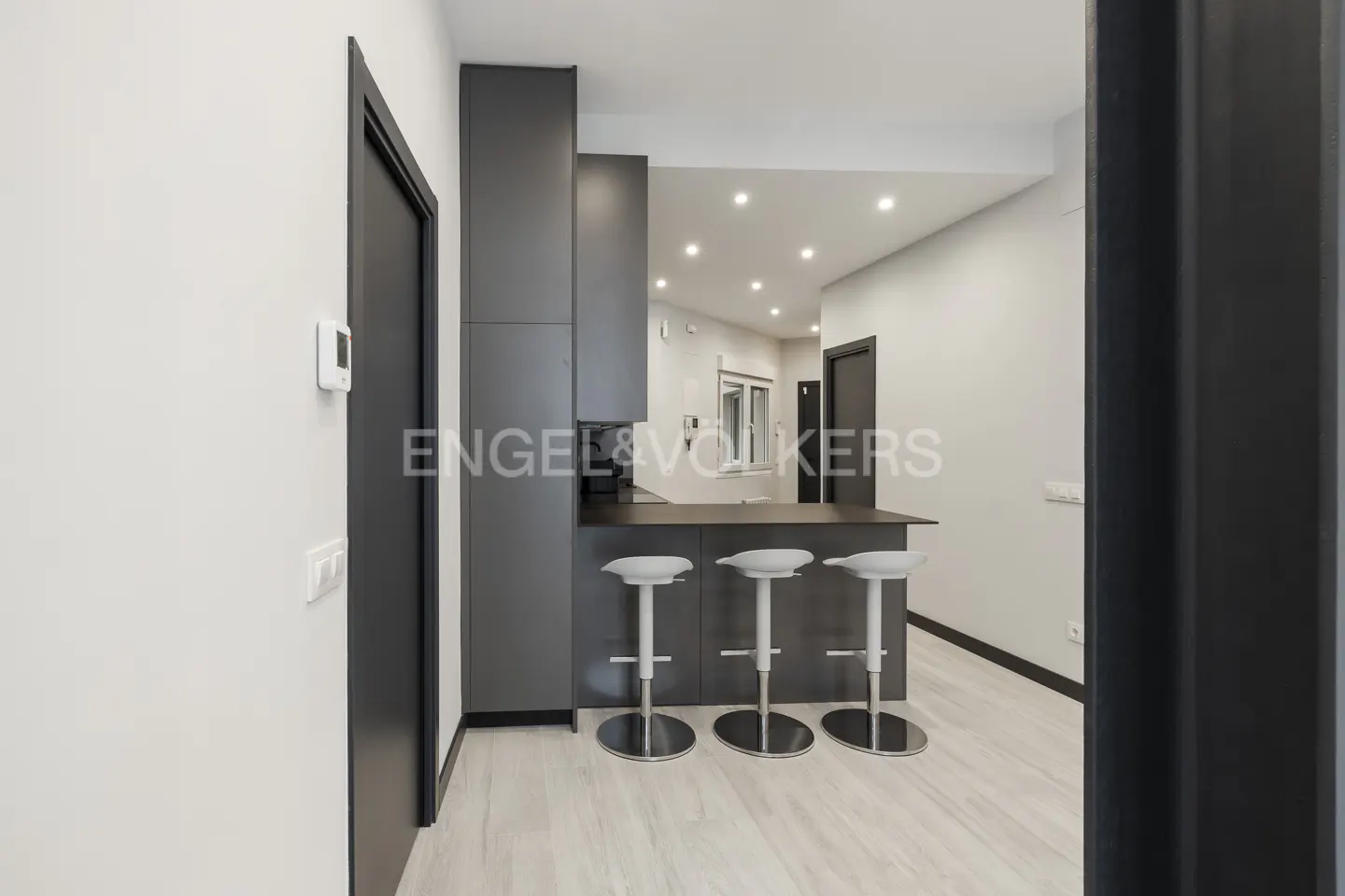 Modern kitchen with gray cabinets, a dark countertop bar, and three white bar stools on a light wood floor.