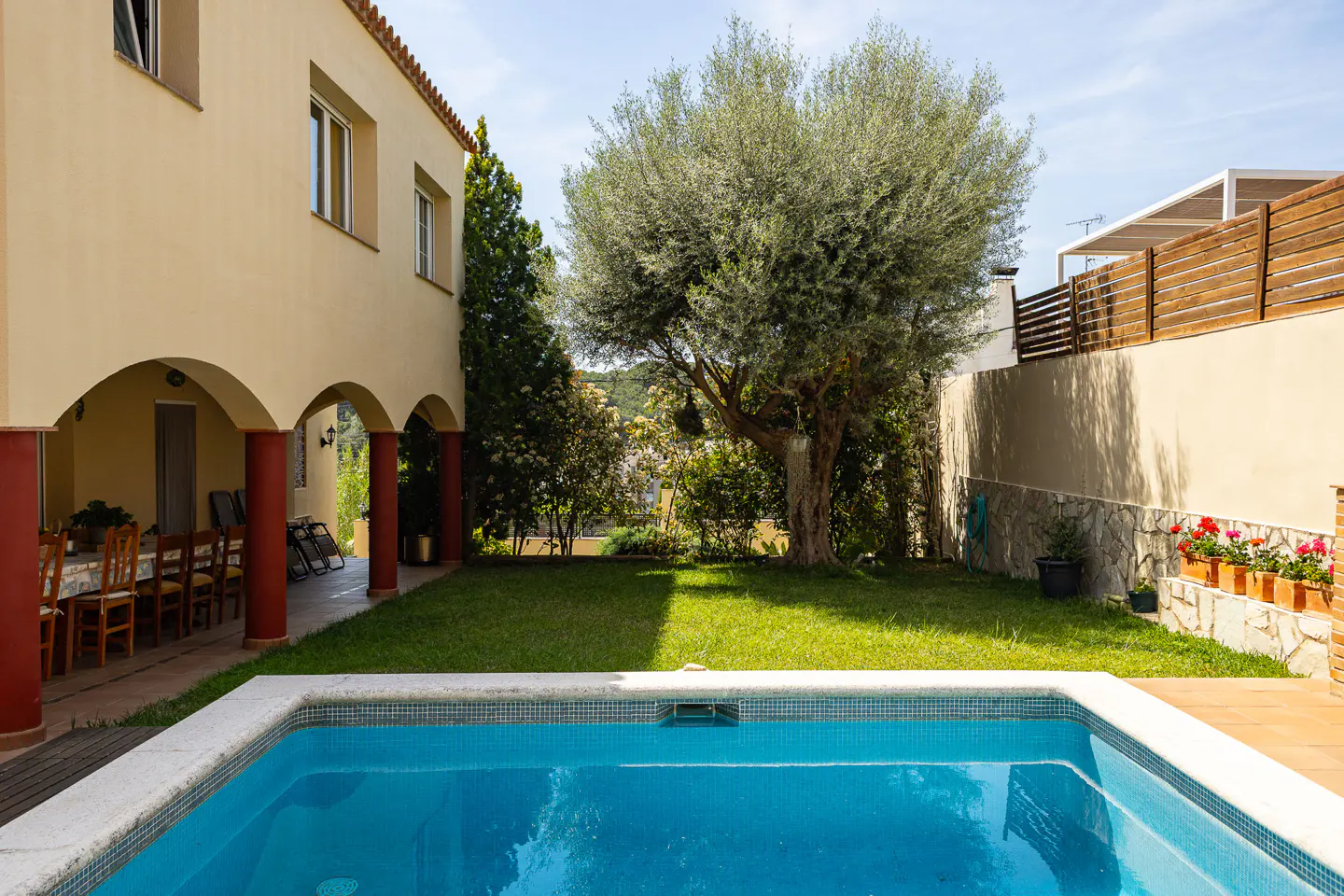 Backyard view of a tan house with red columns, a blue tiled pool, green grass, and a large tree.