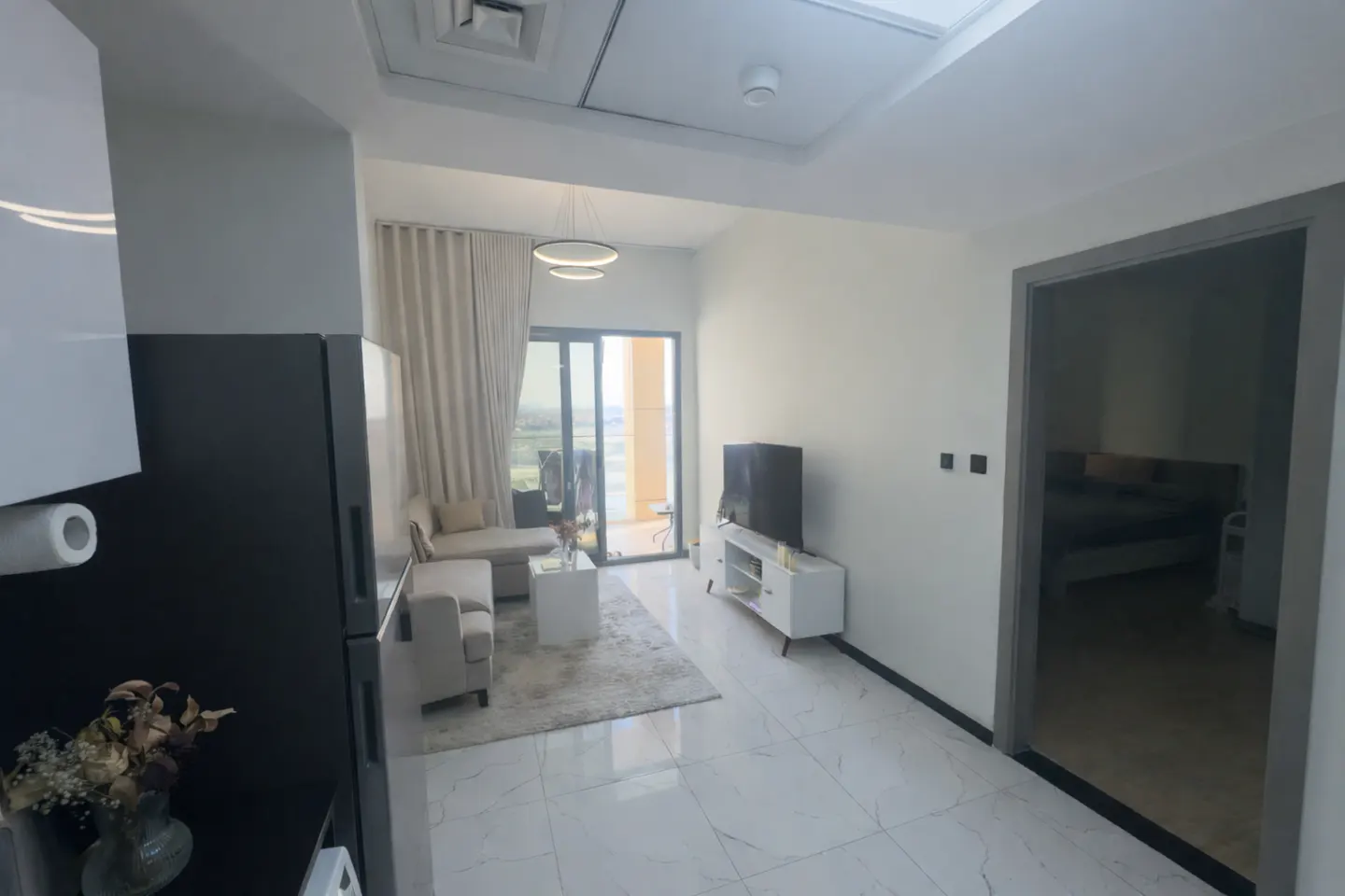 A bright, modern apartment interior with white marble floors, a living room with a beige sofa, and a balcony view.