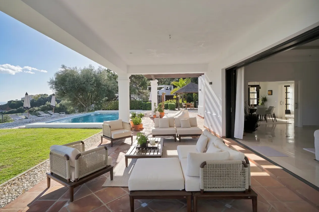 Covered patio with white sofas and chairs on a terracotta tile floor. A pool and green lawn are visible in the background.