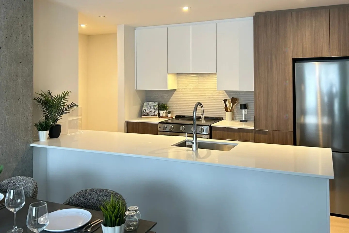 A modern kitchen with white cabinets, stainless steel appliances, and a white countertop island. A dining table with place settings is in the foreground.