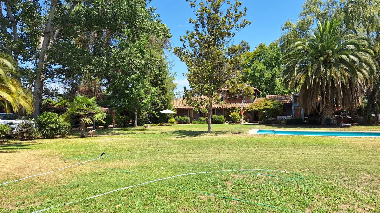 A large, grassy backyard with a swimming pool, mature trees, and a single-story house in the background under a blue sky.