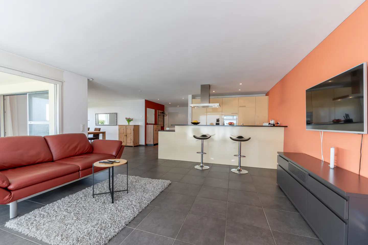 Open-concept living space with a red leather sofa, gray rug, and kitchen island with bar stools. A TV is mounted on an orange wall.