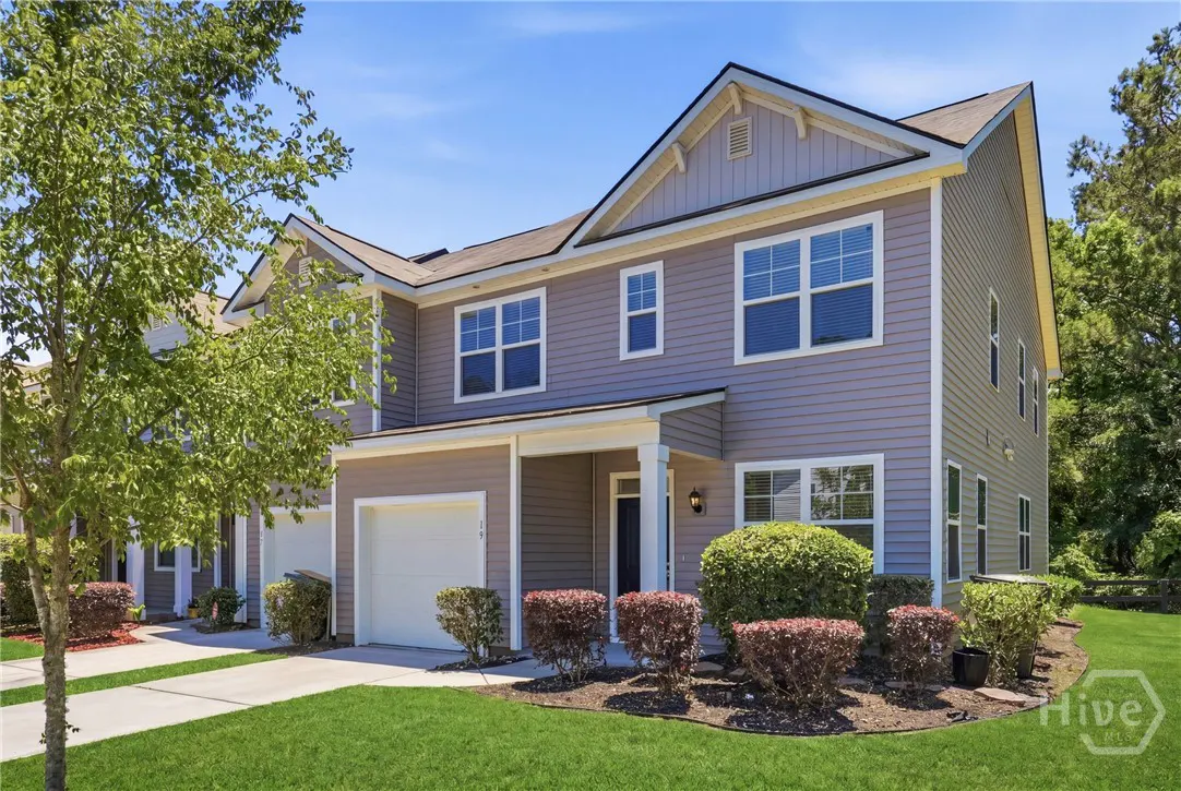 Exterior view of a two-story, gray townhouse with a white garage door and a blue front door. Green lawn and trees surround the building.