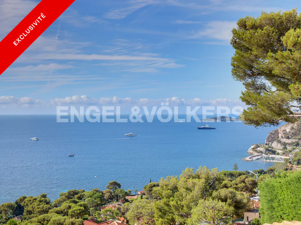 Scenic view of the blue Mediterranean Sea with yachts, lush green trees, and a coastal town under a clear sunny sky.