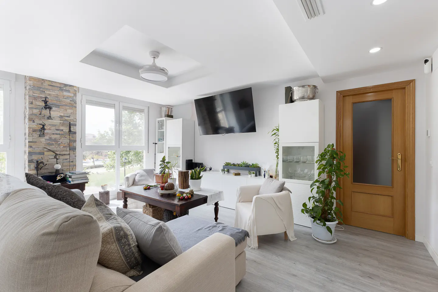 Bright living room with a beige sofa, white armchair, dark wood coffee table, and a large TV mounted on the wall.