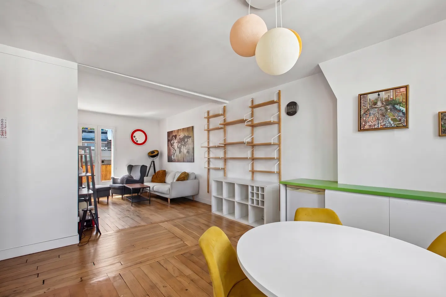 Bright, open-concept living space with hardwood floors, white walls, and modern decor. Yellow chairs surround a white table. Shelving unit and artwork add character.