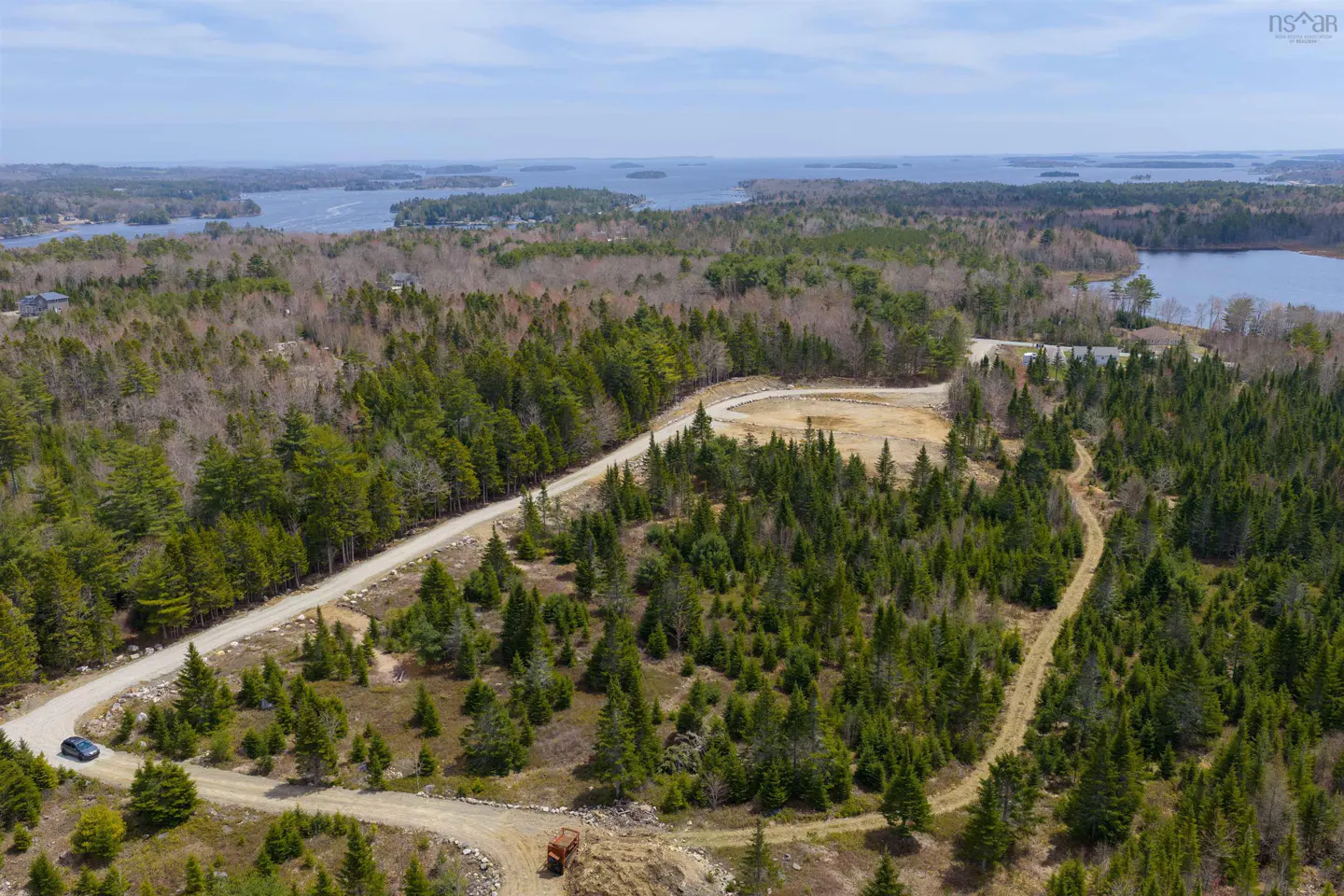 Aerial view of a wooded lot with a dirt road winding through green trees. A blue car is parked near the road, and a lake is visible in the background.