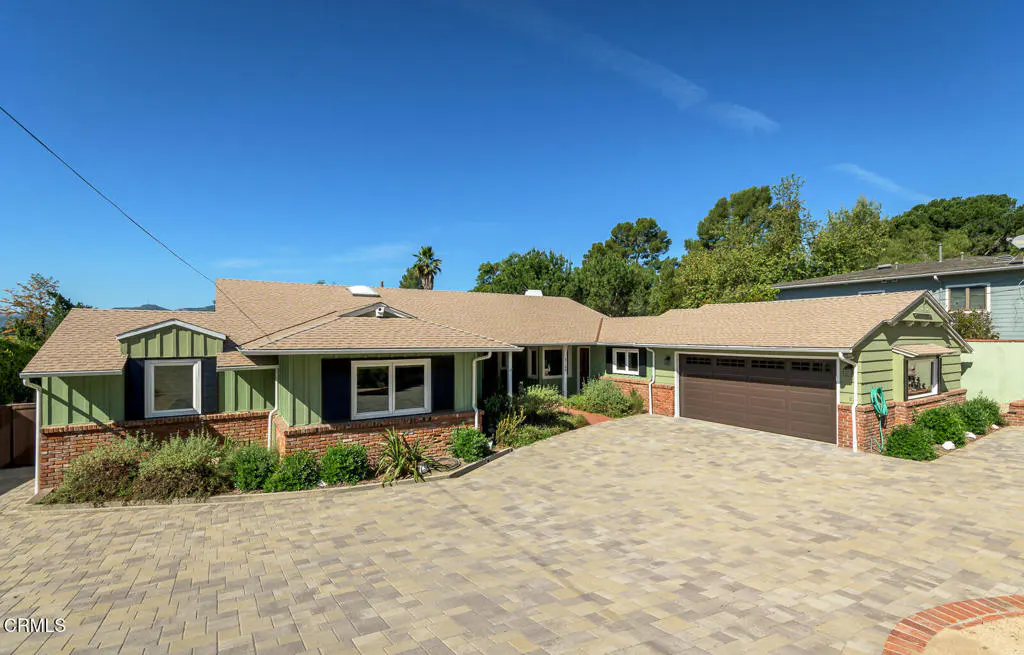 Exterior view of a one-story green house with a brown roof and a brick foundation. A large driveway is in front of the house.
