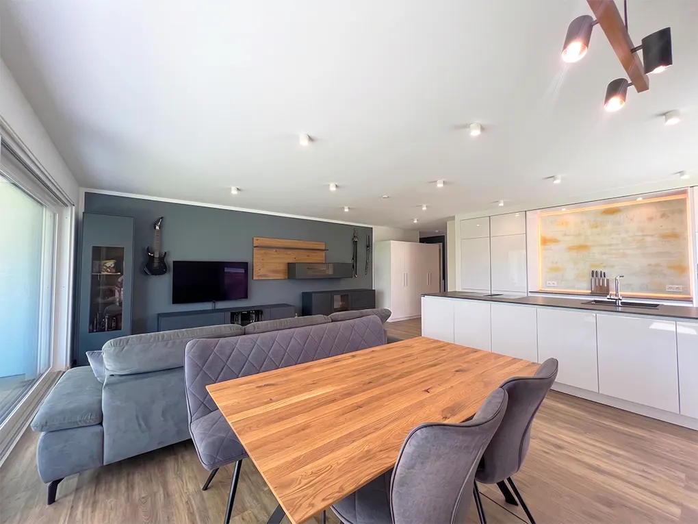 Open-concept living space with a wood dining table, gray sofa, and white kitchen cabinets. A guitar hangs on the gray wall.