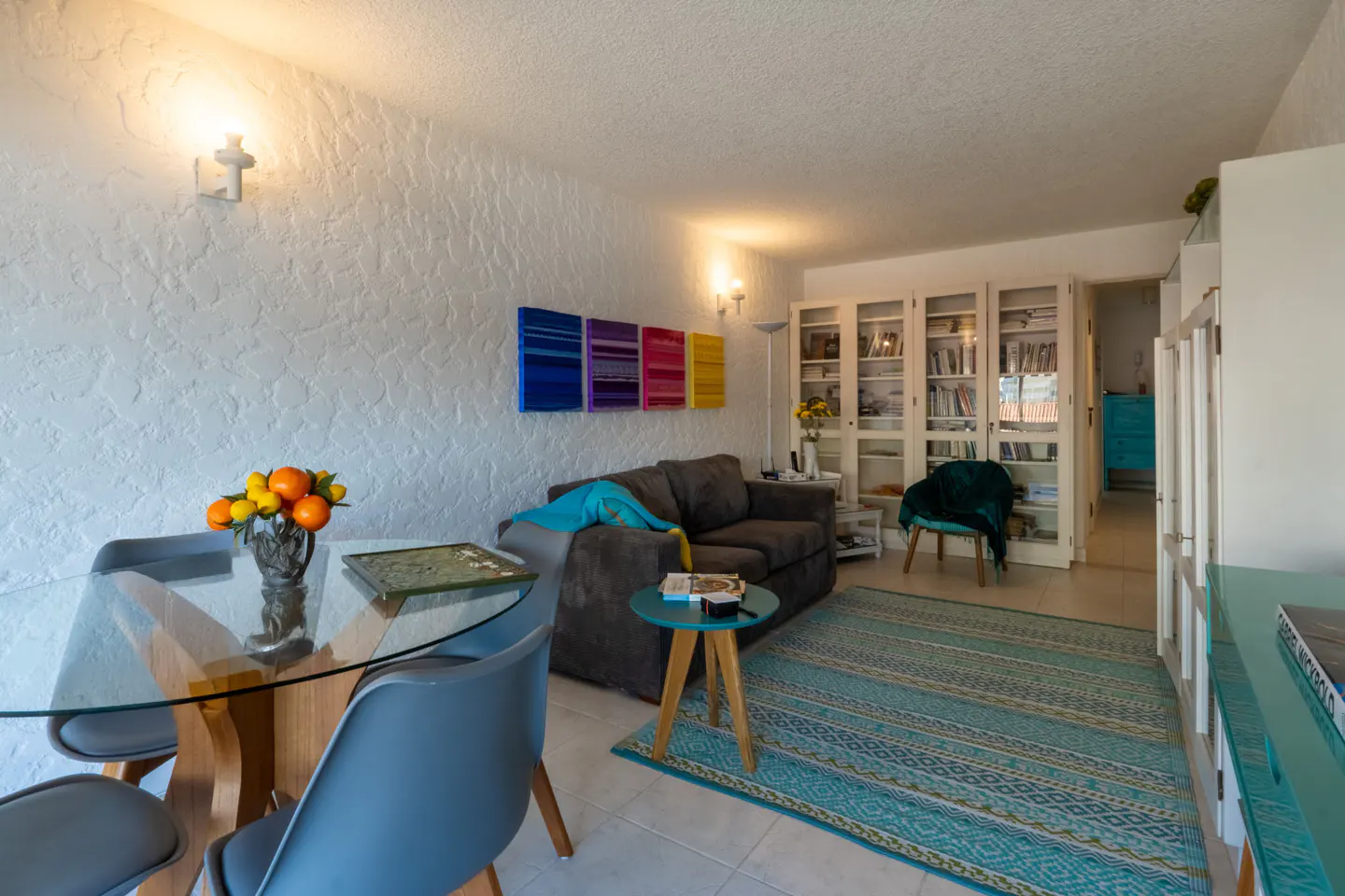 Living room with a glass table, gray chairs, a gray sofa, and colorful artwork on a textured white wall. A blue rug covers the floor.