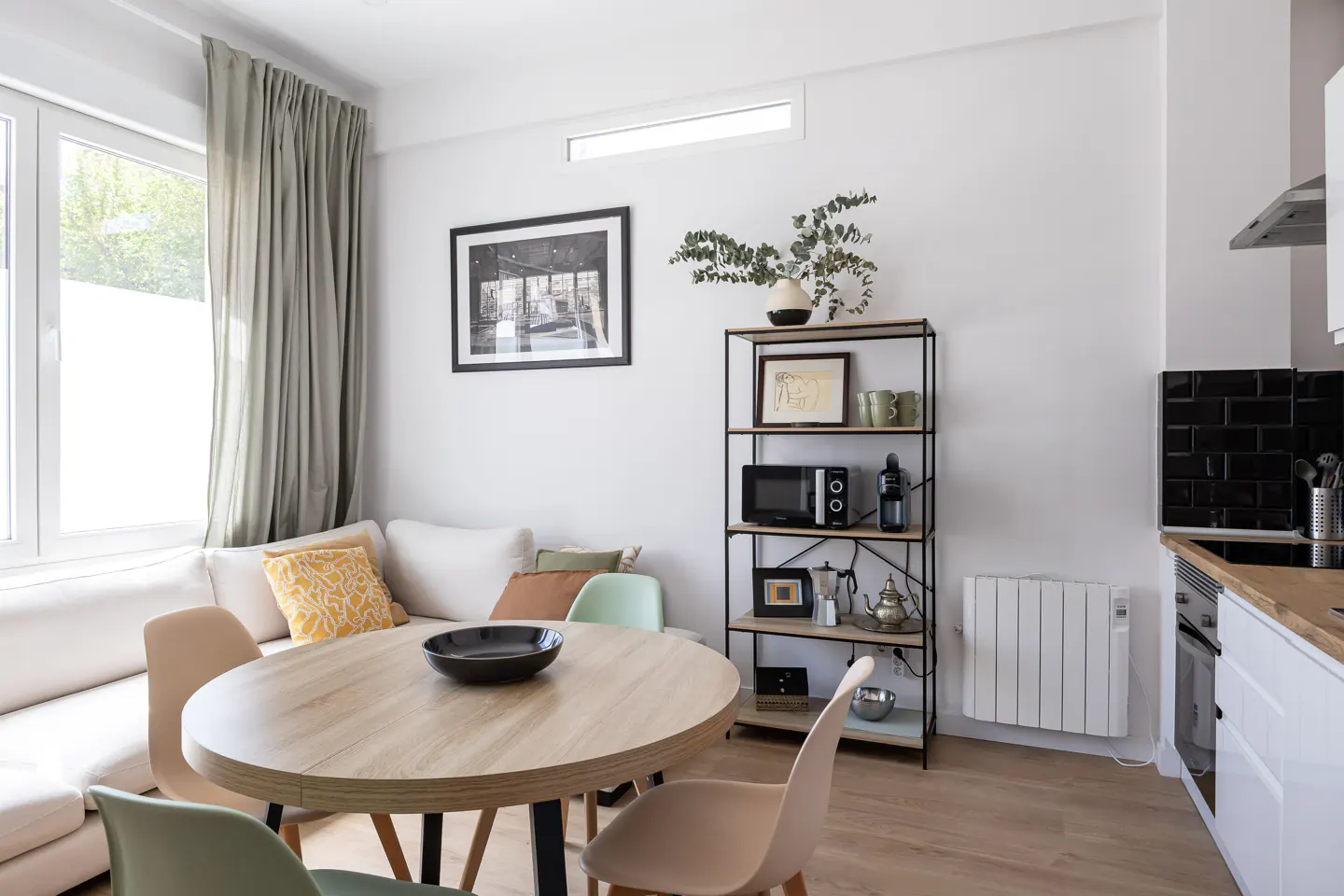 Bright apartment interior with a round table, sofa, and shelving unit. A black bowl sits on the table. The kitchen area is visible in the background.