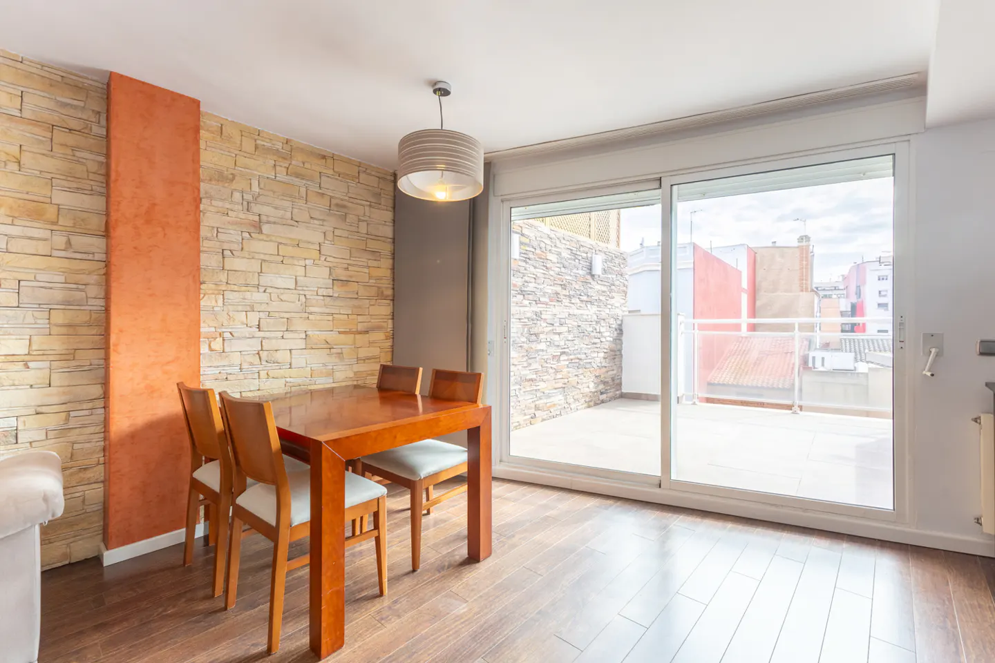 A dining room with a wooden table, four chairs, and a sliding glass door to a balcony. The wall is stone with an orange accent.