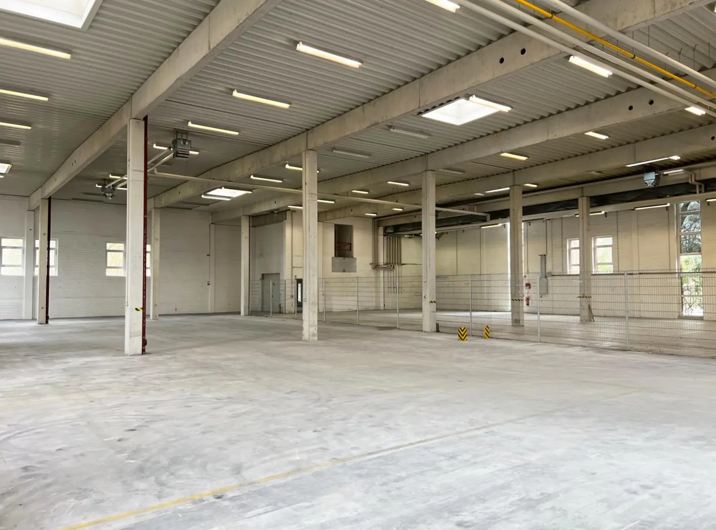 Empty warehouse with concrete floors, white walls, and a high ceiling with fluorescent lights. Concrete pillars support the roof.
