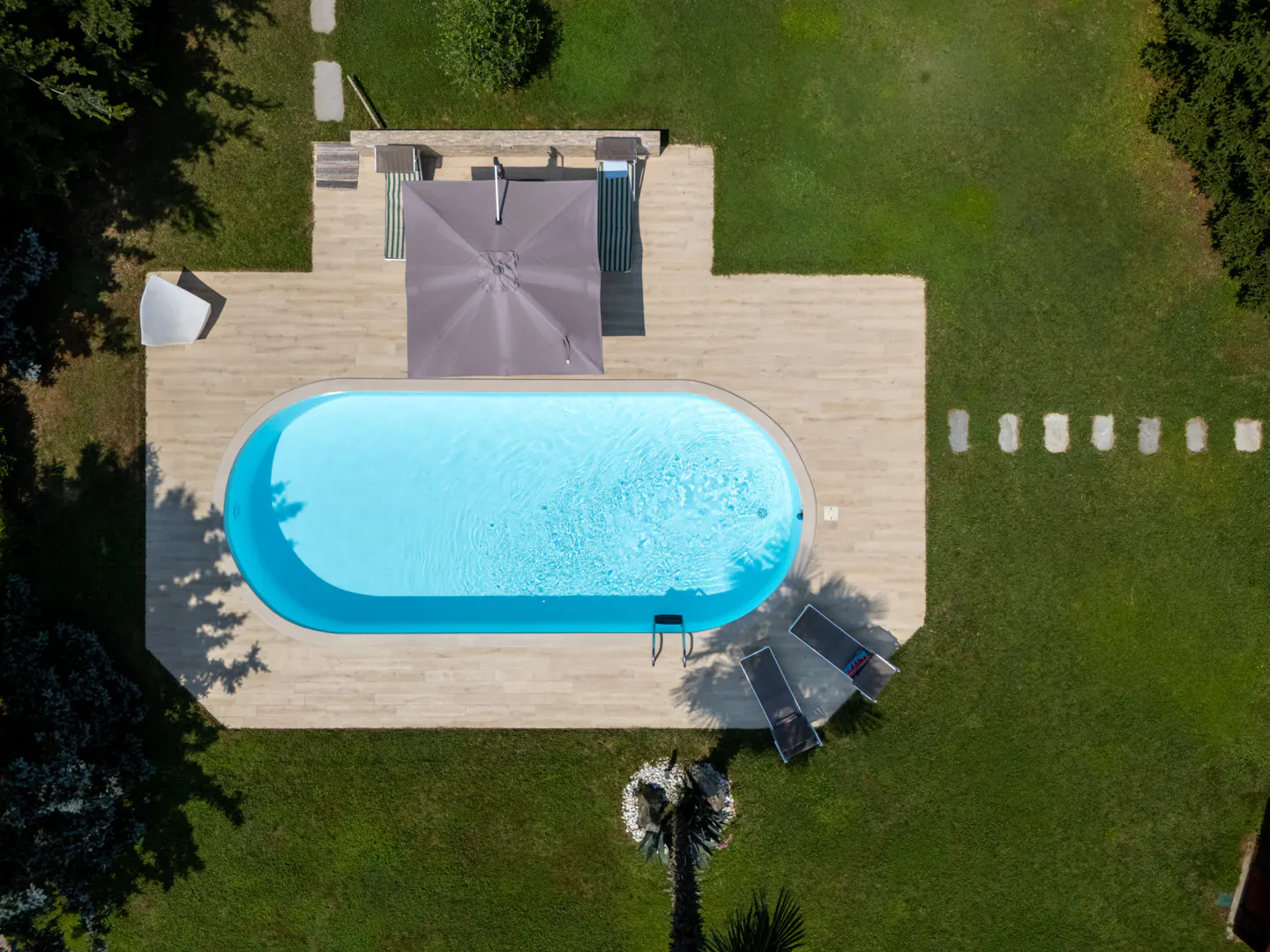 Aerial view of an oval swimming pool with a grey umbrella, lounge chairs, and stone patio set in a lush green lawn.