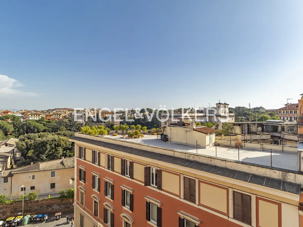 View of Rome cityscape with terracotta buildings, green trees, and blue sky. Engel & Völkers logo is superimposed on the image.