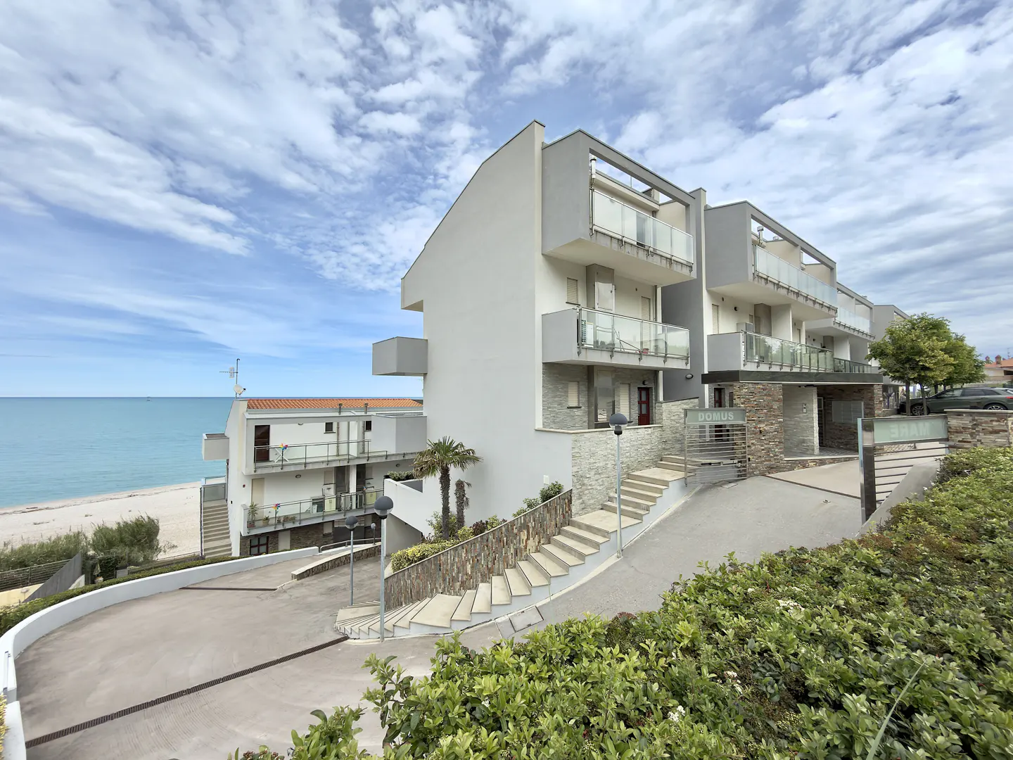 Exterior view of modern white apartments with glass balconies, near a sandy beach and blue sea under a cloudy sky.