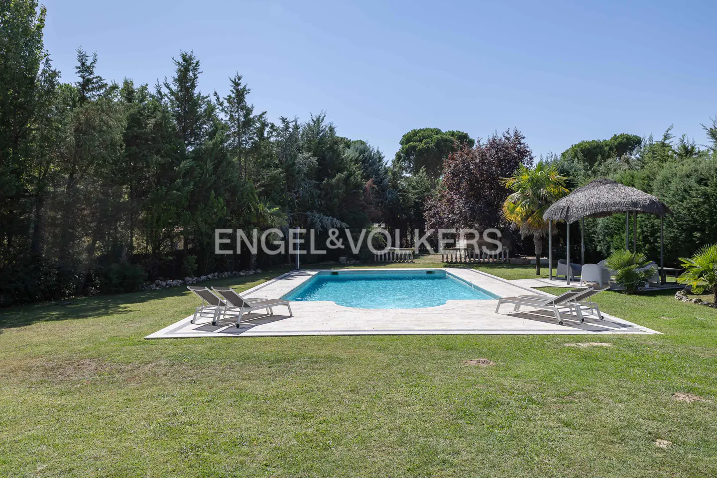 A backyard with a pool, lounge chairs, a gazebo, and green grass. Trees surround the yard.