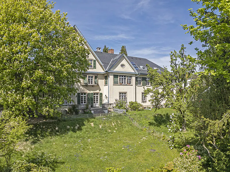 A large, cream-colored house with green shutters, a dark roof, and a grassy lawn. Trees surround the house.