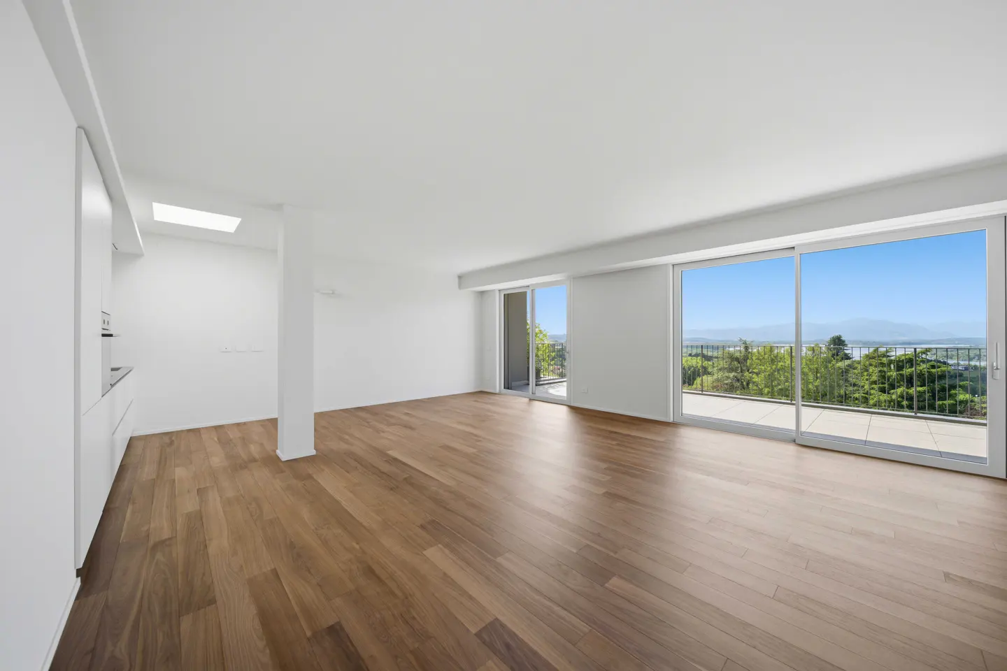 Bright, empty room with hardwood floors, white walls, and sliding glass doors to a balcony with a view of trees and a lake.