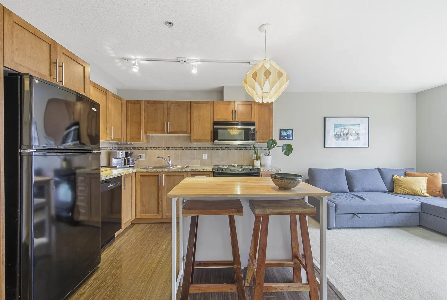 Open-concept living space with wood floors, featuring a kitchen with wood cabinets, black appliances, and a blue sofa.
