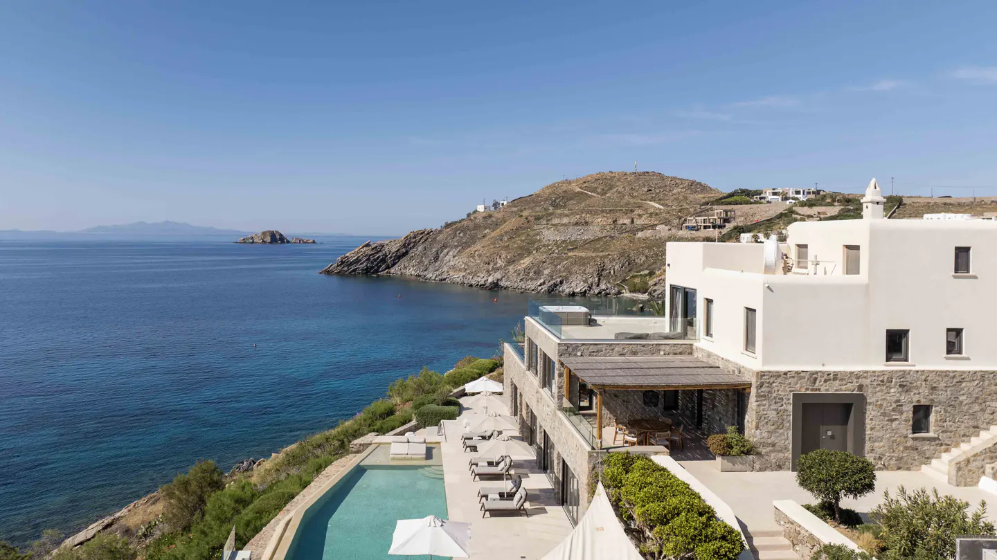 Luxury villa with pool overlooking the Aegean Sea. White building with stone accents, lounge chairs, and blue water.