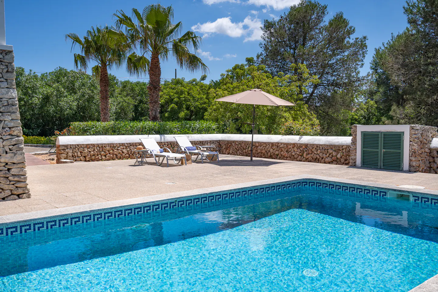 A turquoise pool with Greek key tiles, two white lounge chairs, and a beige umbrella on a sunny patio.