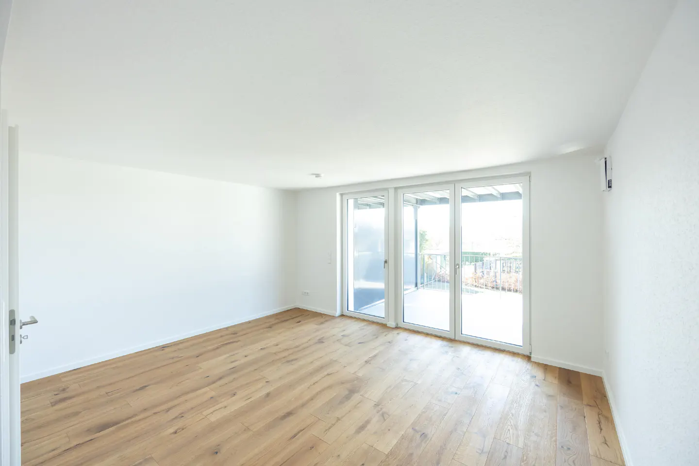 Bright, empty room with light wood floors and white walls. Sliding glass doors lead to a balcony.