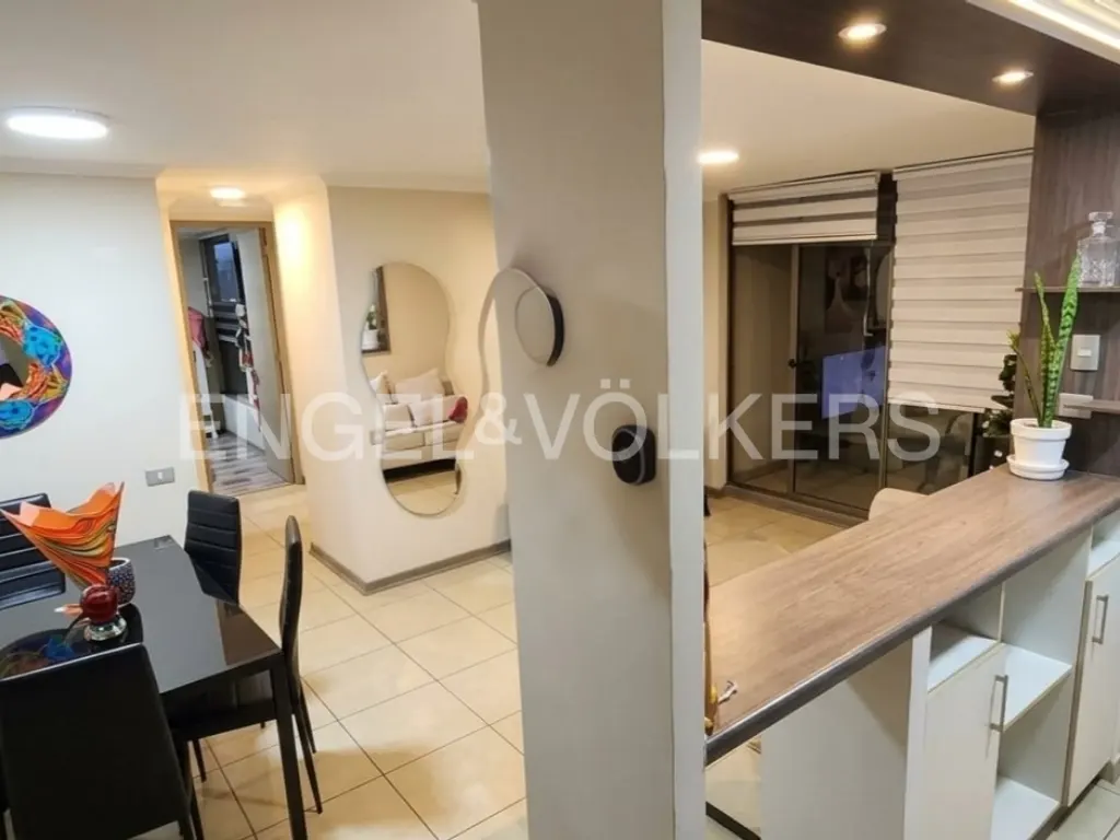 A modern apartment interior with a black dining table, a kitchen counter, and a living room visible through a doorway.