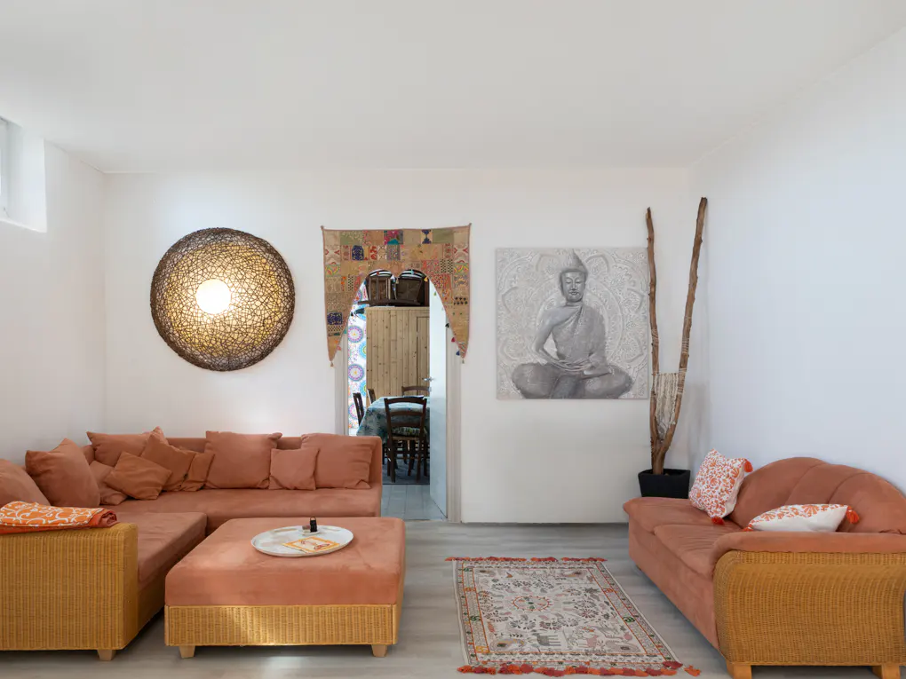 Living room with peach sofas, wicker accents, and a Buddha painting. A woven light fixture and colorful doorway add unique touches.