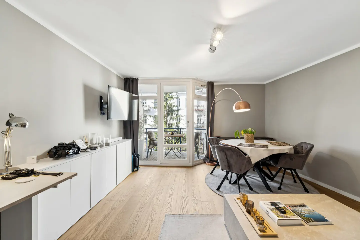 A modern living room with gray walls, light wood floors, and a balcony. A dining table with gray chairs sits near the balcony doors.
