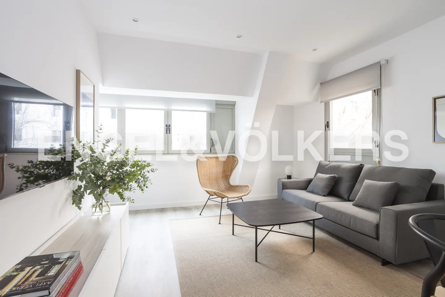 Bright living room with gray sofa, black coffee table, and wicker chair on a beige rug. White walls and a TV on a white console.