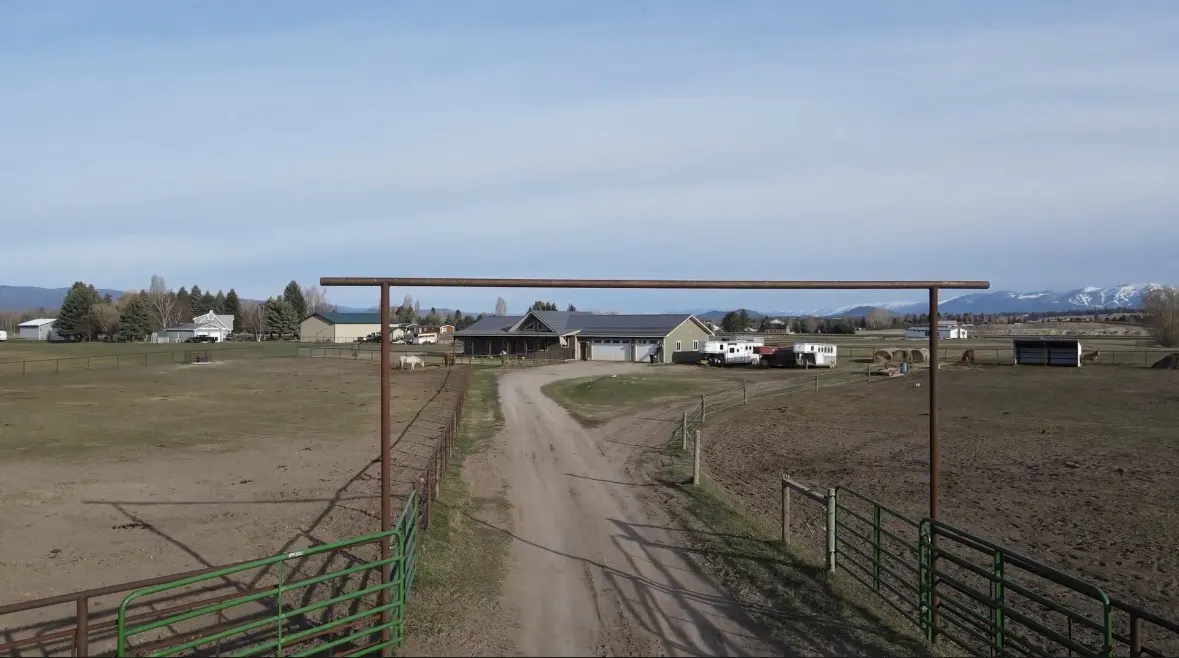 View of a ranch through a metal gate, with a dirt road leading to a house and barn. Mountains are visible in the background.
