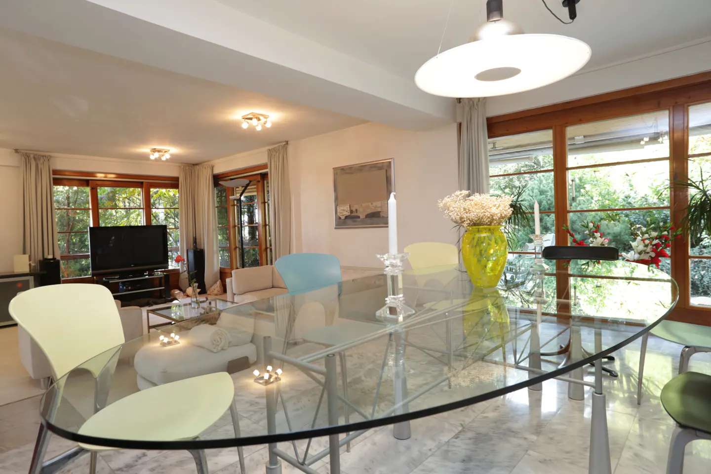 Bright living and dining area with a glass table, colorful chairs, and large windows overlooking a green garden.