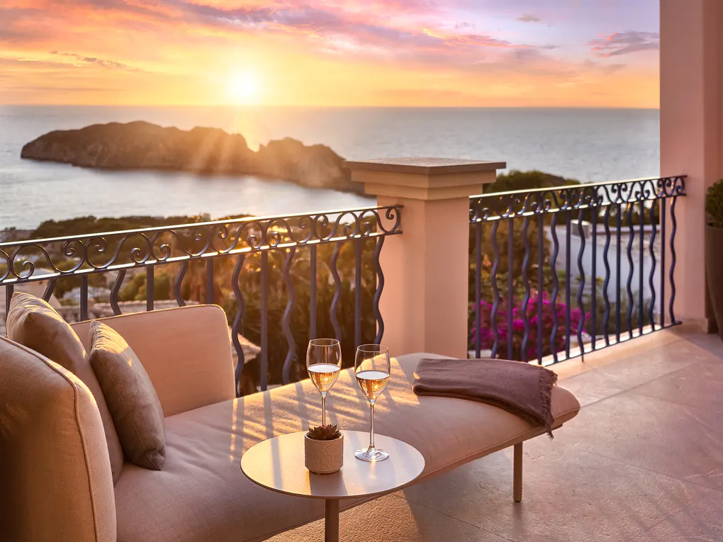 Balcony view at sunset with two wine glasses on a table next to a beige lounge chair overlooking the ocean.