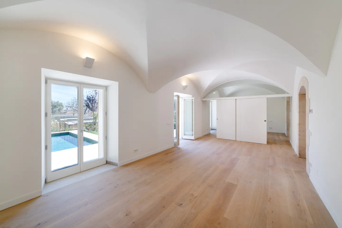 Bright, empty room with white walls, arched ceiling, and light wood floors. French doors open to a pool. White sliding doors lead to another room.