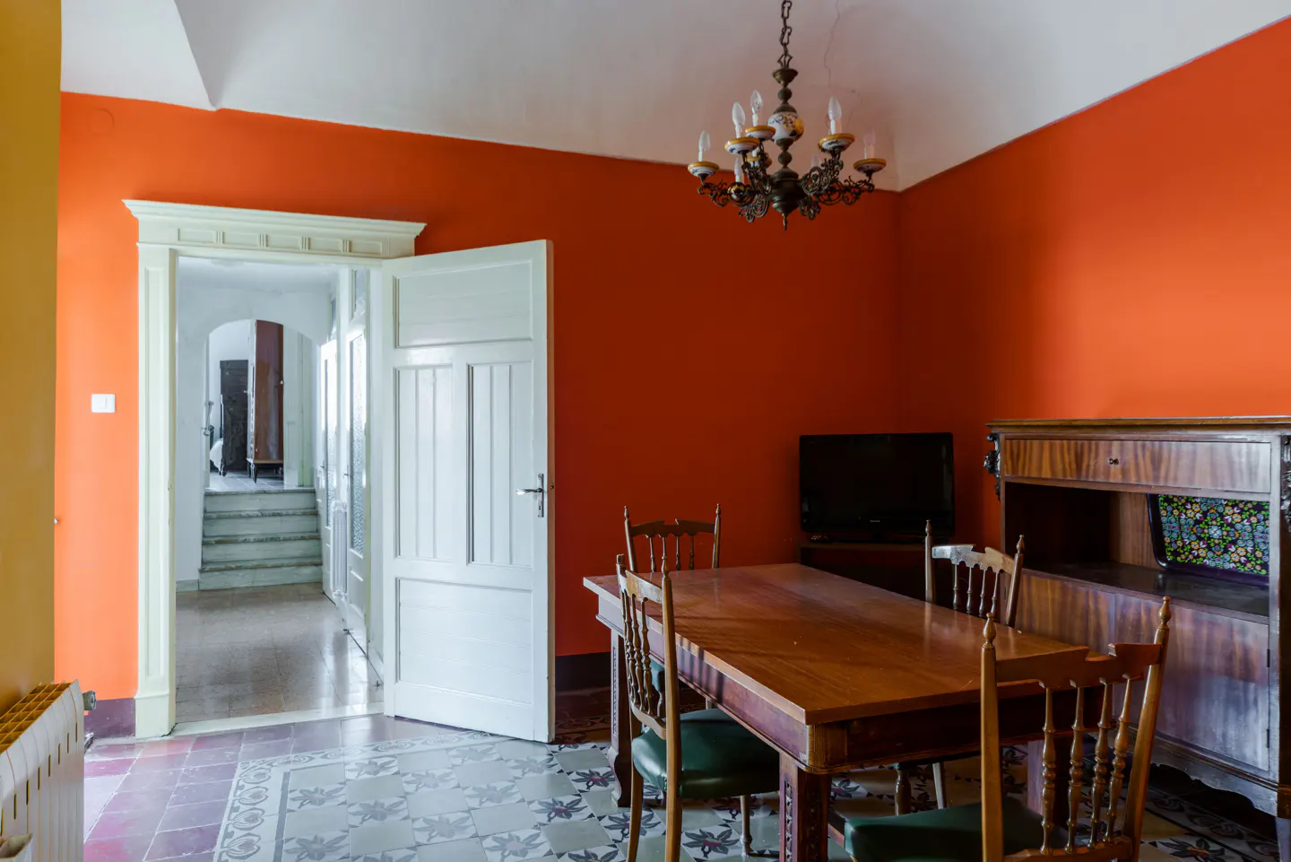 A dining room with orange walls, a wooden table and chairs, and a vintage chandelier. A doorway leads to another room with stairs.