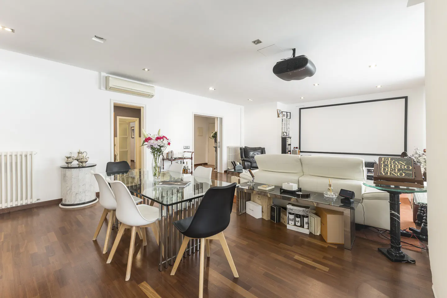 Bright living room with a glass table, white chairs, and a projector screen. Hardwood floors and white walls create a modern space.