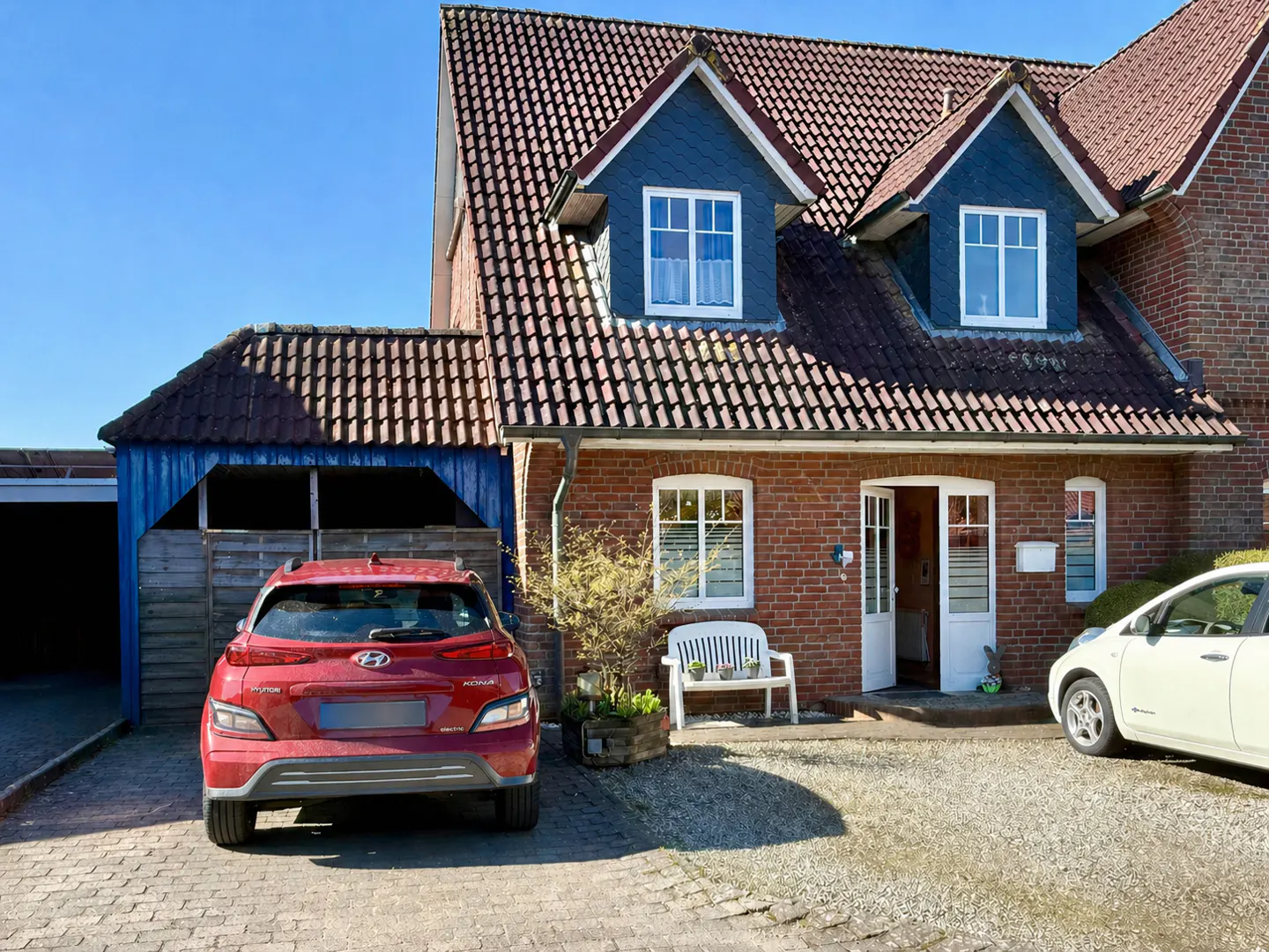 A two-story brick house with a red tile roof, dormers, and two cars parked in front.