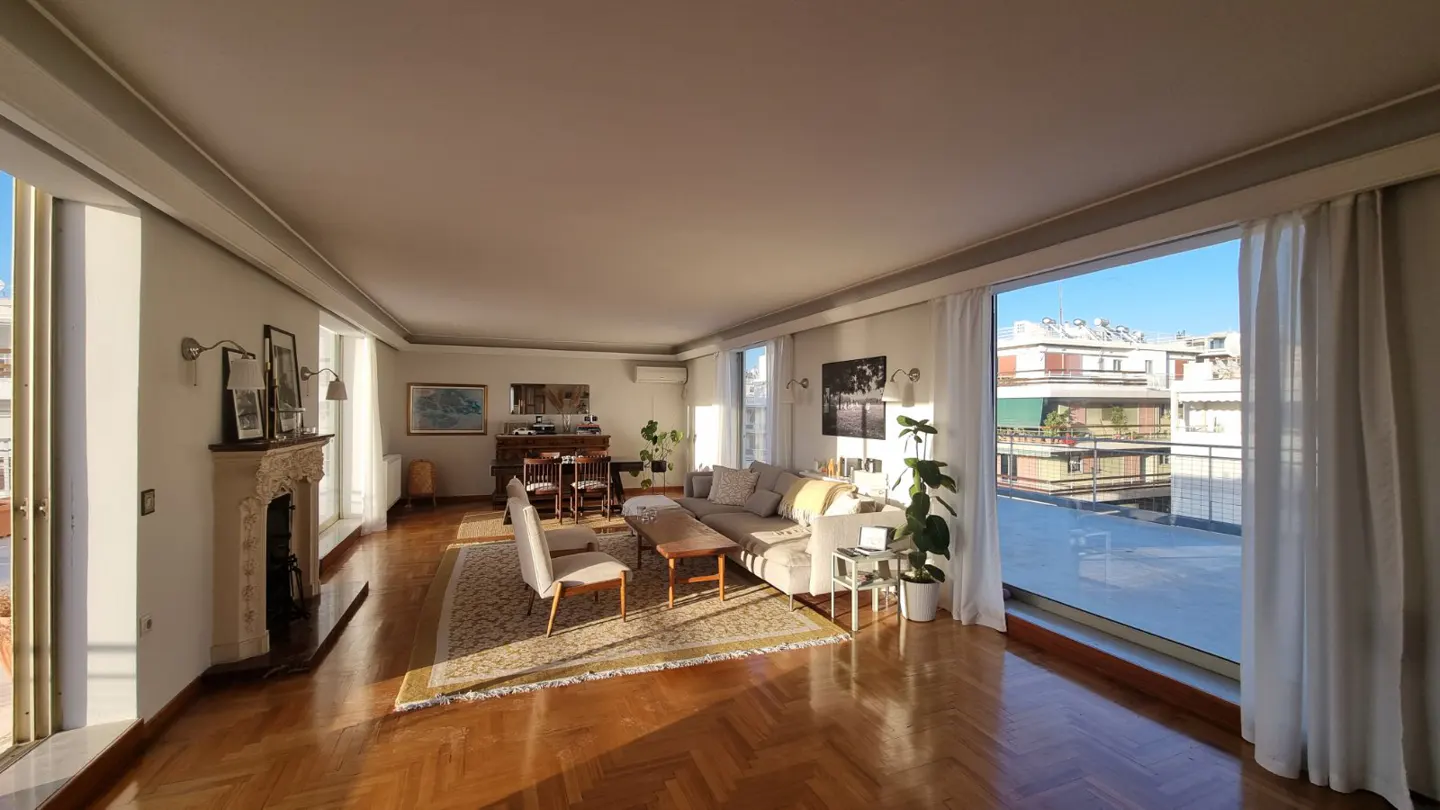 Bright, spacious living room with hardwood floors, a fireplace, and large windows overlooking a city view. A sofa, chairs, and a rug furnish the space.