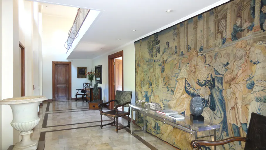 Hallway with marble floors, white walls, and a large tapestry. A silver table and chairs are visible.