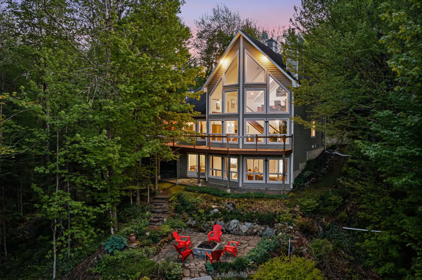 A multi-story home with large windows and a deck, set in a lush forest with a stone patio and red chairs at dusk.