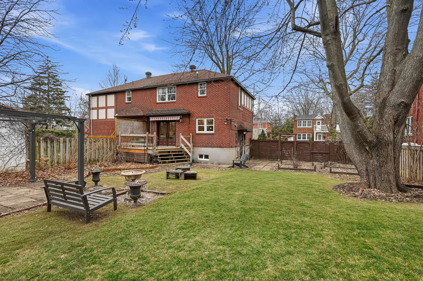 Backyard view of a two-story red brick house with a green lawn, wooden deck, bench, and bare trees under a blue sky.