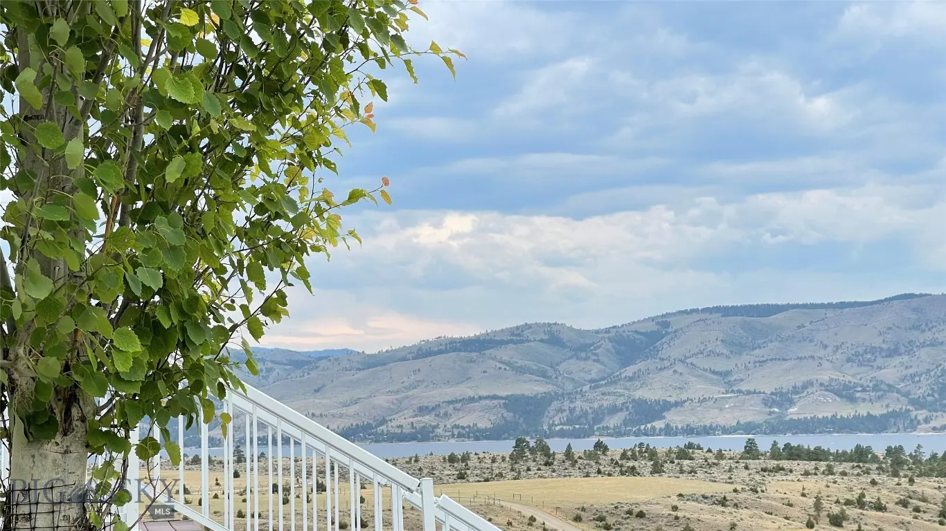 View from a deck with white railings, a tree with green leaves, and a mountain and lake view under a cloudy sky.