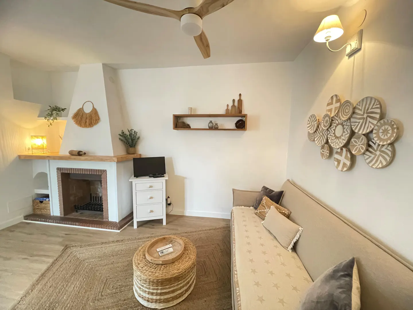 Living room with white walls, a fireplace, a beige sofa with pillows, and a round straw table on a jute rug. A wooden ceiling fan is visible.