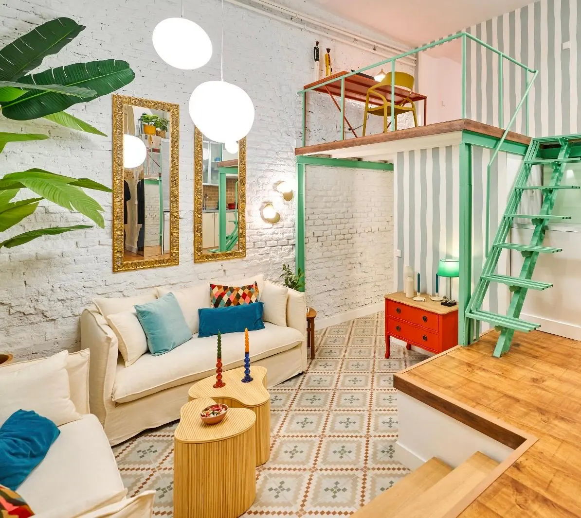 Bright living room with white brick walls, patterned tile floor, and a loft with green metal frame and stairs. Cream sofa with blue pillows.
