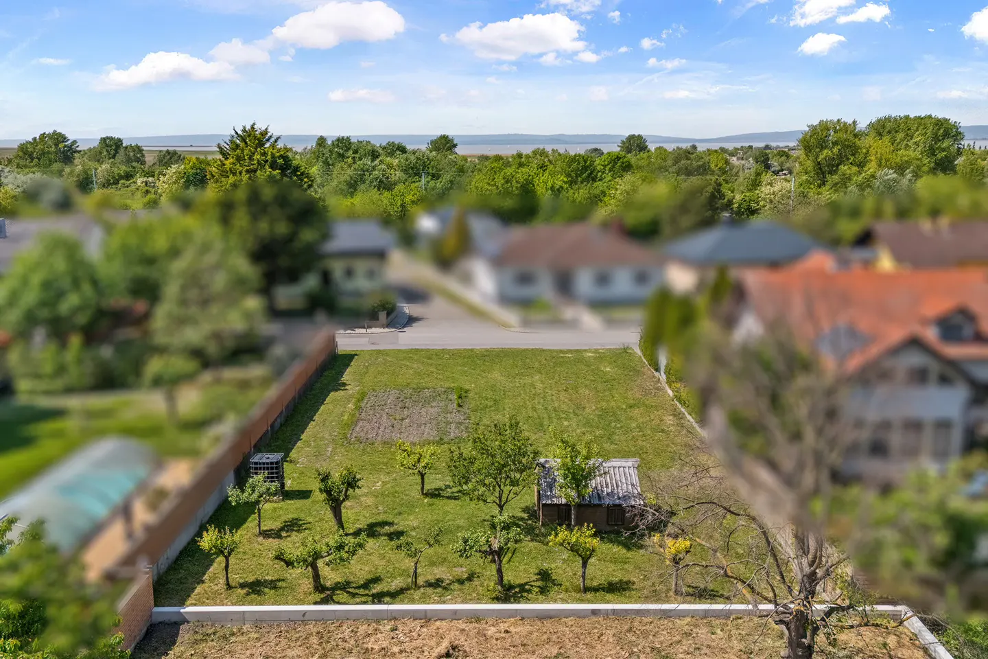 Aerial view of a grassy lot with small trees, a shed, and a distant view of a lake under a blue sky.