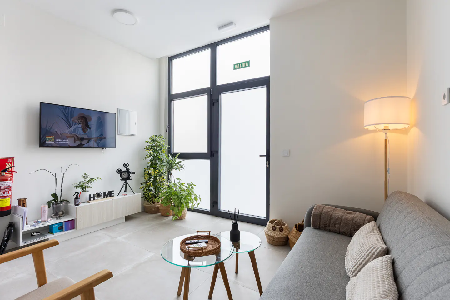 Living room with gray sofa, glass table, TV, plants, and frosted glass doors with an exit sign.