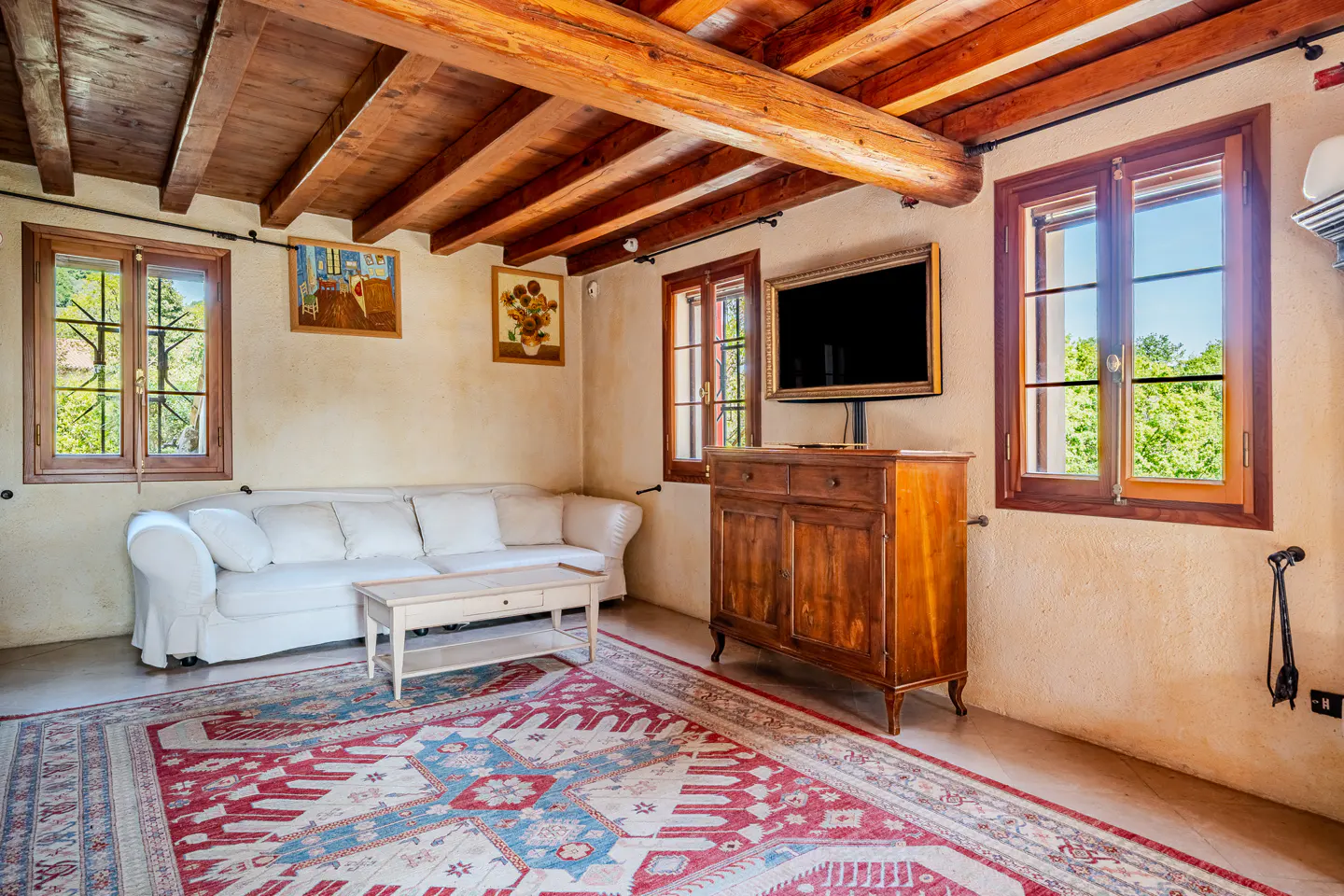 Living room with exposed wood beams, white sofa, coffee table, and a red and blue patterned rug. A wooden cabinet with a TV sits against the wall.