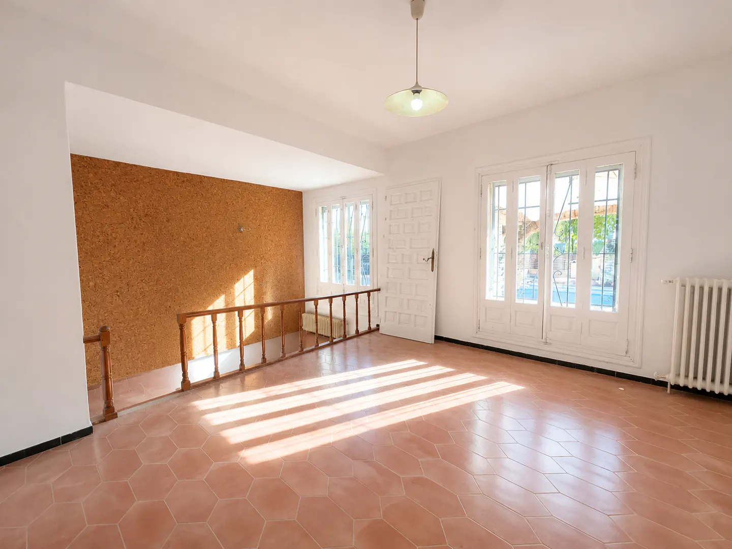 Bright, empty room with terracotta tile floor, white walls, and a cork accent wall. Sunlight streams through the windows.